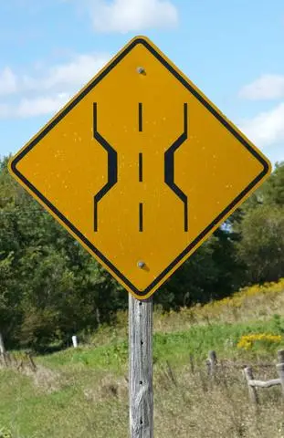 Yellow diamond sign showing two lines pinching inward, indicating the road narrows