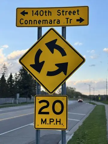 Yellow diamond roundabout sign with circular arrows and a 20 MPH plaque, under a 140th Street and Connemara Trail street sign