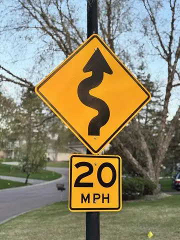 Yellow diamond sign with a winding arrow above a 20 MPH plaque in a residential street